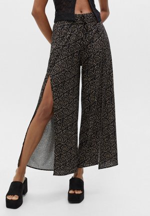 Femme portant un pantalon large noir à motifs floraux avec de grandes fentes sur les côtés, associé à des sandales plateformes noires à bout ouvert.