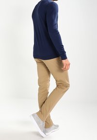 Námořnicky modrý pletený svetr s dlouhými rukávy, v kombinaci s lehce béžovými chinos kalhotami a šedými vysokými teniskami s bílými podrážkami, viděno z bočního úhlu.