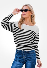 Mujer con cabello rubio que lleva gafas de sol estilo gato negro, suéter a rayas blancas y negras, jeans azules y pendientes de aro dorados.