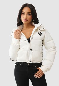 Biała kurtka puchowa o skróconym kroju, czarne guziki i detal z logo. Żebrowane mankiety i dół oraz boczne kieszenie. W połączeniu z czarnymi dżinsami.