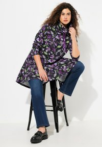 Túnica negra con un patrón floral púrpura y verde, cuello drapeado y mangas largas, combinada con jeans azules y mocasines negros.