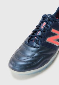 Zapato de fútbol de cuero azul marino con paneles texturizados, acentos en rosa y una suela translúcida. Presenta un destacado logo "N" y cordones seguros.