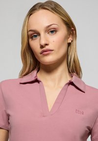 Kortärmad pikétröja i ljusrosa tyg. Har krage, v-ringad design och en broderad "POLO CLUB" logotyp på vänster bröst.