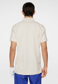 Beige kortärmad polo med krage, slät textur och diskret logotyp på ryggen. Bärs med blå byxor och en vit handske.