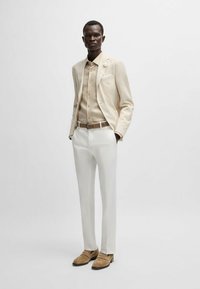 Light beige tailored jacket with a pocket square, paired with a beige button-up shirt and white trousers. Suede loafers complete the look.