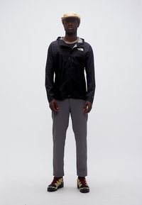 Άνδρας που φοράει μαύρο μπουφάν North Face, γκρι παντελόνι, μπεζ καπέλο και μαύρα-κίτρινα ορειβατικά παπούτσια, στέκεται μπροστά σε λευκό φόντο.