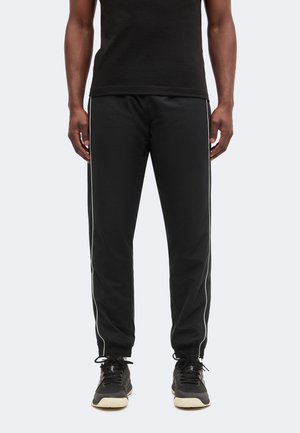 Hombre con pantalones de chándal negros con rayas blancas en los lados, zapatillas negras y camiseta negra, de pie frente a un fondo liso.