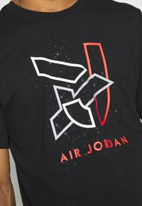Černé tričko s stylizovaným logem Air Jordan v bílé a červené barvě. Design zahrnuje geometrické tvary a tečkované pozadí.