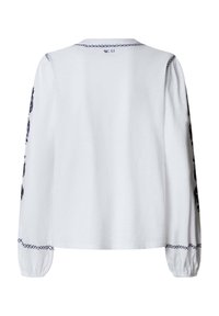 Blouse blanche à manches longues avec des motifs en points de croix noirs le long du col, des poignets et des manches, étiquetée "W.11" dans la nuque au dos.