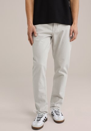 Persoon draagt lichtgrijze slim-fit jeans met witte sneakers met zwarte strepen en witte veters, staand tegen een effen achtergrond.