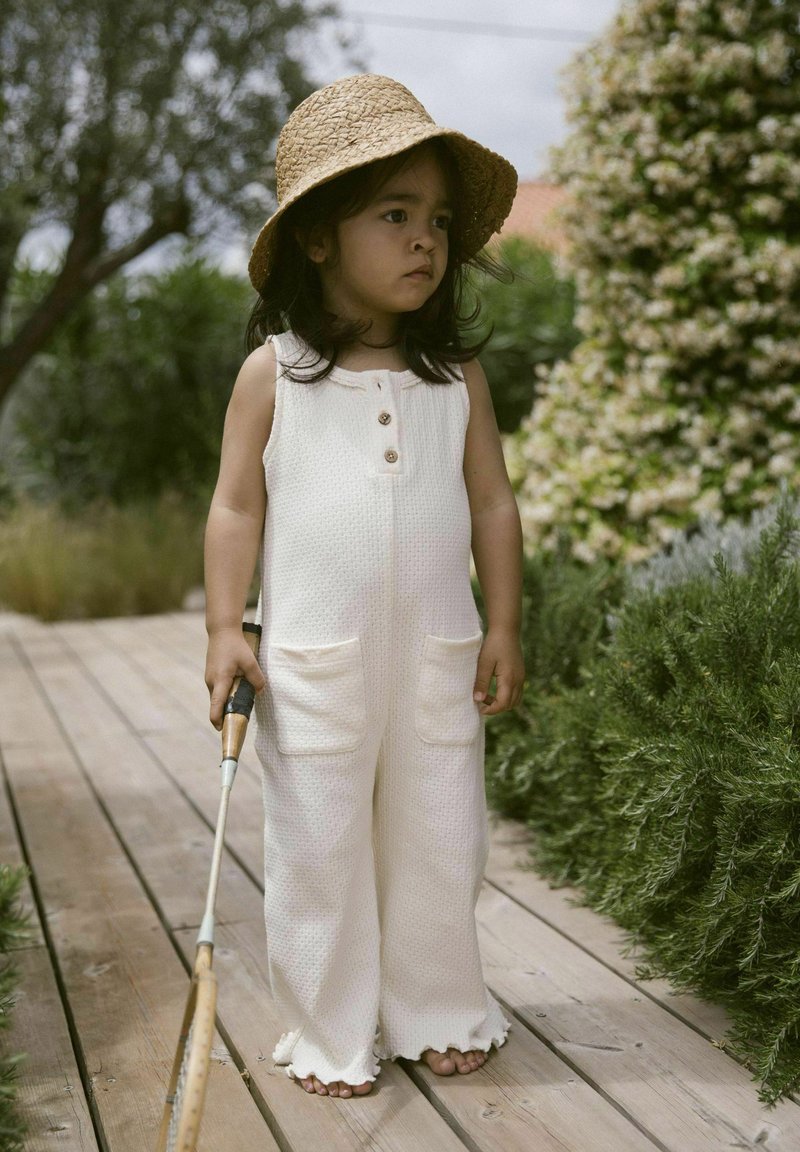Salopette en coton texturé beige clair avec deux poches avant et des boutons. Portée avec un chapeau en paille, un enfant se tient sur un chemin en bois à côté de verdure.