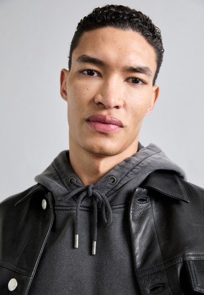 Czarna skórzana kurtka na szarym hoodies z sznurkami. Zbliżenie na twarz męskiego modela, ukazujące teksturowaną skórę i krótkie kręcone włosy.
