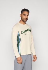 Camisola de manga comprida bege claro com a inscrição "Cream City" em verde, com riscas laterais verdes e azuis. Tecido macio e design de corte solto.