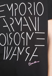 Černé tričko s výrazným bílým grafickým textem v moderním písmu, s malým růžovým logem v pravém dolním rohu. Materiál: bavlna.