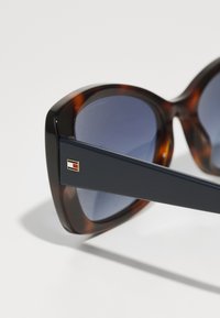 Ochelari de soare tip cat-eye din tortoishell, cu brațe de culoare albastru închis. Lentilele au un gradient închis. Prezintă un accent mic cu logo pe braț.