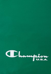 Tela verde con una superficie texturada que presenta un prominente logo blanco de "Champion" y el texto "U.S.A.", ubicado en la esquina inferior derecha.