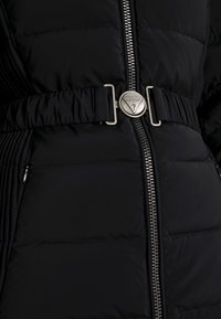 Doudoune noire avec une fermeture éclair métallique et une ceinture avec un logo en métal argenté circulaire. Tissu texturé avec un motif matelassé.