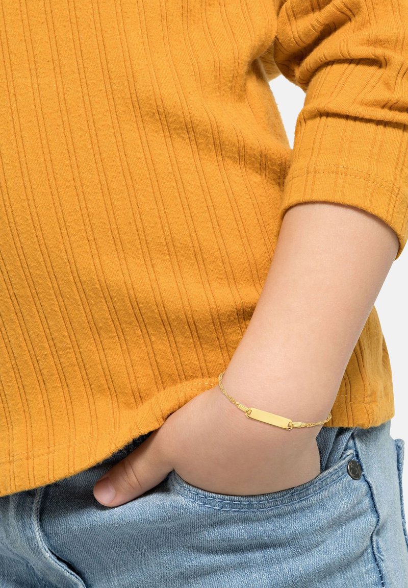 Niño con una camiseta de manga larga de canalé mostaza y vaqueros azules, con una pulsera rectangular de oro en la muñeca descansando en el bolsillo.