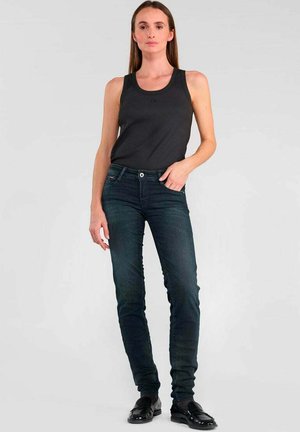 Un débardeur noir sans manches associé à un jean skinny bleu foncé. La tenue présente une texture lisse et un design minimaliste. Des mocassins noirs complètent le look.