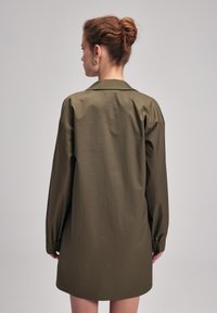 Manteau léger à manches longues, vert olive, avec une coupe décontractée, doté d'un col et de poignets élastiques. Texture lisse avec un minimum de détails.