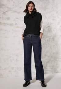 Sort striktrøje med cowl-neck og uden ærmer over en hvid langærmet skjorte, parret med mørkeblå lige ben jeans og sorte ankelstøvler.