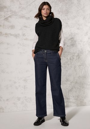 Top sin mangas de punto negro con cuello tipo cowl sobre una camisa blanca de manga larga, combinado con jeans de pierna recta azul oscuro y botas negras hasta el tobillo.