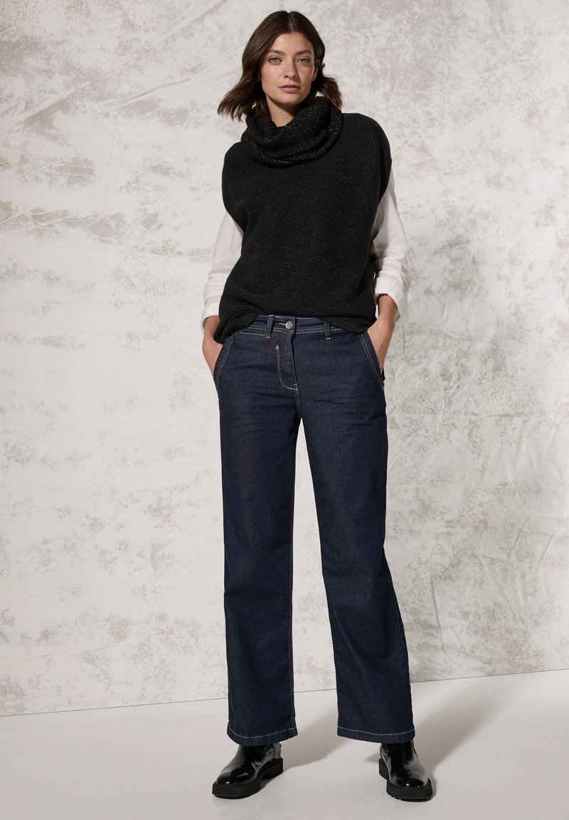 Sort striktrøje med cowl-neck og uden ærmer over en hvid langærmet skjorte, parret med mørkeblå lige ben jeans og sorte ankelstøvler.