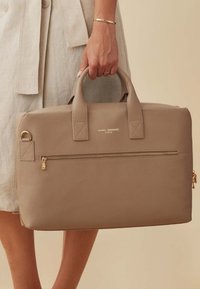 Beige leren schoudertas met dubbele handvatten, een voorzak met rits en gouden hardware-accenten. Textuurafwerking met zichtbaar merklogo.