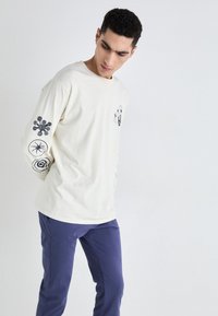 Cămașă crem cu mâneci lungi și desene grafice negre pe mâneci, asortată cu pantaloni bleumarin. Cămașa are o croială lejeră și o textură moale.