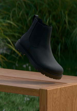 Schwarzer Leder-Knöchel-Stiefel mit elastischen Seiteneinsätzen, der über einer Holzbank im Freien schwebt, mit unscharfem grünem Pflanzenhintergrund.