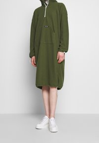 Olivgrön huvdklänning med dragkedjefunktion vid halsen, framficka och dragsko i nederkant. Matchas med vita sneakers med tjock sula.