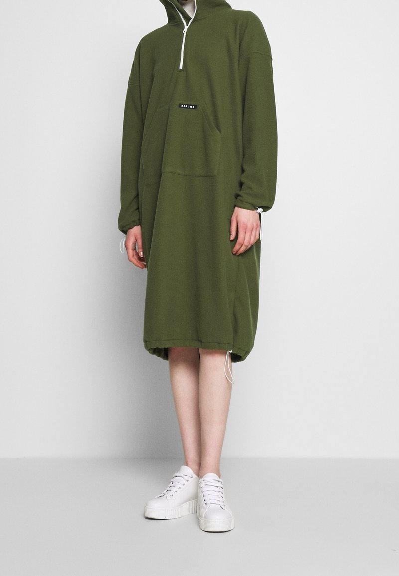 Olivgrön huvdklänning med dragkedjefunktion vid halsen, framficka och dragsko i nederkant. Matchas med vita sneakers med tjock sula.