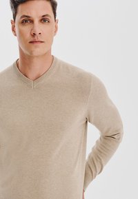 Beżowy sweter w serek z miękkiego, teksturowanego dzianiny. Dopasowany krój z długimi rękawami, z delikatnym prążkowanym wykończeniem dekoltu i mankietów.