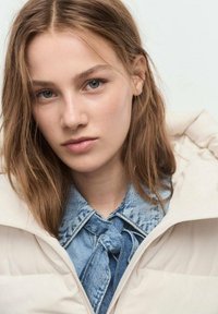 Chaqueta de plumas beige claro con cuello alto, llevada sobre una camisa de mezclilla azul con un detalle de lazo atado. Maquillaje natural y cabello hasta los hombros.