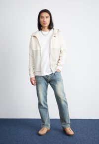 Lys farget hettegenser med glidelås og kontrasterende mørkere paneler, båret over en hvit t-skjorte, kombinert med falmede blå jeans og beige sko.