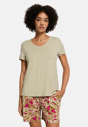Vrouw met krullend haar, gekleed in een beige T-shirt met V-hals en beige korte broek met roze en bruine abstracte patronen, staand met één hand in de zak.