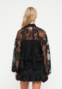 Sort gennemsigtig bluse i blonde med blomstermønster og flæser, med lange ærmer og høj krave, stylet over en sort flæset nederdel.