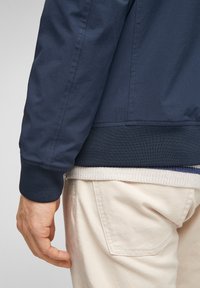 Veste bomber bleu marine à texture lisse, avec poignets et ceinture côtelés, portée sur un pull beige clair et un pantalon crème.