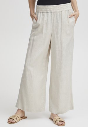 Pantalons larges de couleur crème avec une texture côtelée, dotés d'une taille élastique et de poches latérales. Associés à des sandales à glissière beiges.