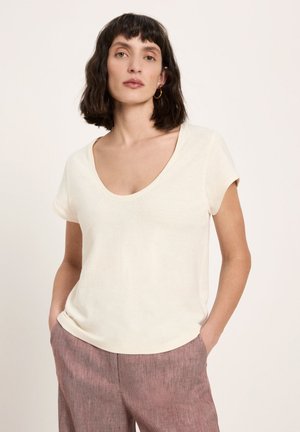 Woman with short dark hair wearing a cream scoop-neck short sleeve top and mauve textured pants, hands in pockets, neutral background.
