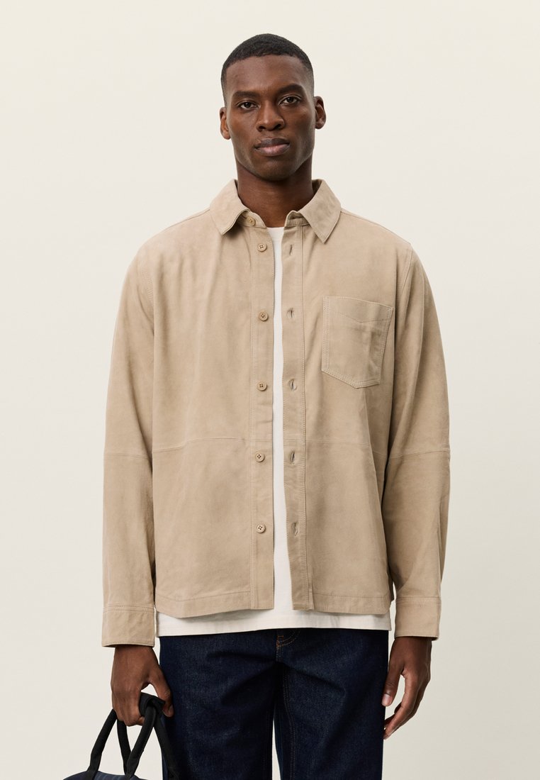 Μπεζ πουκάμισο-σακάκι από σουέντ με εμπρός κλείσιμο με κουμπιά, γιακά και τσέπη στο στήθος, συνδυασμένο με λευκό t-shirt και σκούρο τζιν.