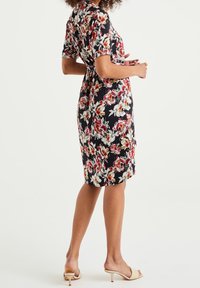 Robe florale à manches courtes, fond noir, fleurs rouges et blanches, forme cintrée, longueur genou, associée à des talons beiges.