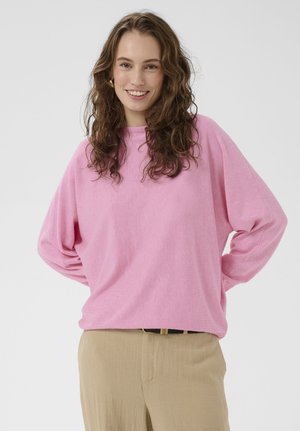 Lachende vrouw met krullend bruin haar draagt een roze trui met lange mouwen en beige broek, poseert met handen op haar heupen tegen een witte achtergrond.