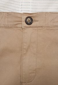 La taille d'un pantalon beige boutonné sur une chemise blanche rayée rentrée, montrant les coutures détaillées et la texture du tissu.