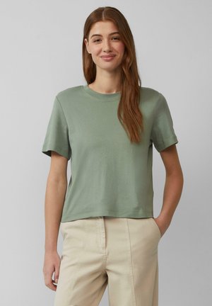 Korte mouwen, lichtgroene katoen T-shirt met een ronde halslijn en een ontspannen pasvorm, gecombineerd met beige broek, tegen een neutrale achtergrond.