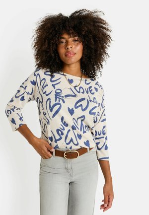 Femme aux cheveux bouclés portant un pull beige avec des motifs en bleu "love" et en forme de cœur, un pantalon gris clair et une ceinture marron, posée avec une main sur la hanche.