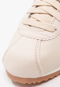 Gros plan sur la pointe d'une basket beige en cuir texturé, lacets beiges, coutures détaillées et semelle en caoutchouc marron clair avec des stries.