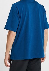 Man som bär en enfärgad blå t-shirt med kort ärm och svarta byxor, sedd bakifrån, stående mot en vit bakgrund.