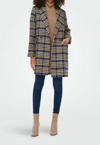 Vrouw in een blauwe en beige geruite jas, beige coltrui, donkerblauwe spijkerbroek en beige enkellaarzen, staande met één hand in haar zak.