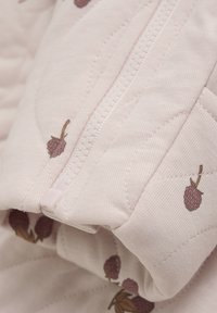 Vadderat tyg i mjuk rosa med små bärmönster. Har en vit dragkedja och detaljerad sömnad som framhäver en texturerad yta.